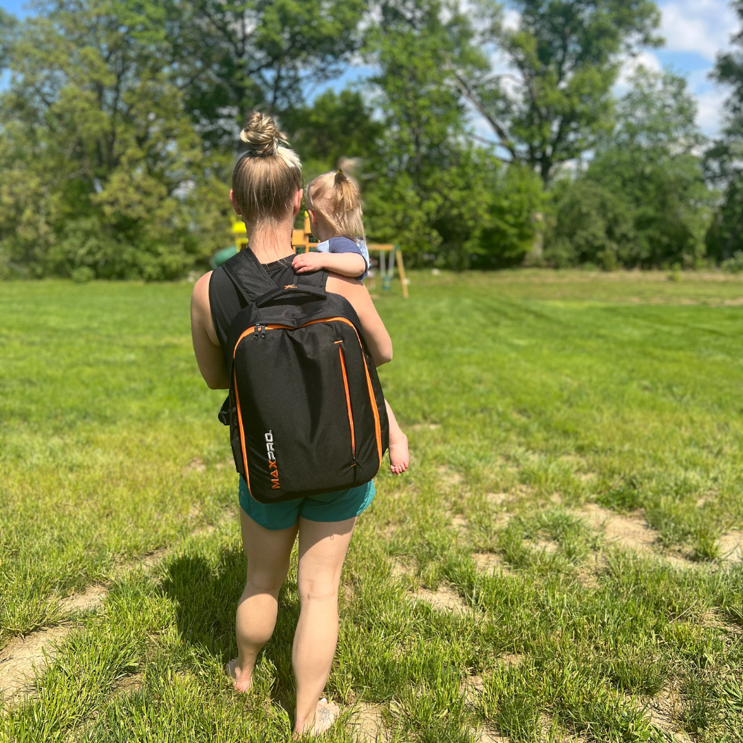 Woman walking in a grassy field holing a child, wearing a maxpro black backpack with orange accents.
