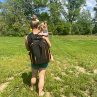 Woman walking in a grassy field holing a child, wearing a maxpro black backpack with orange accents.