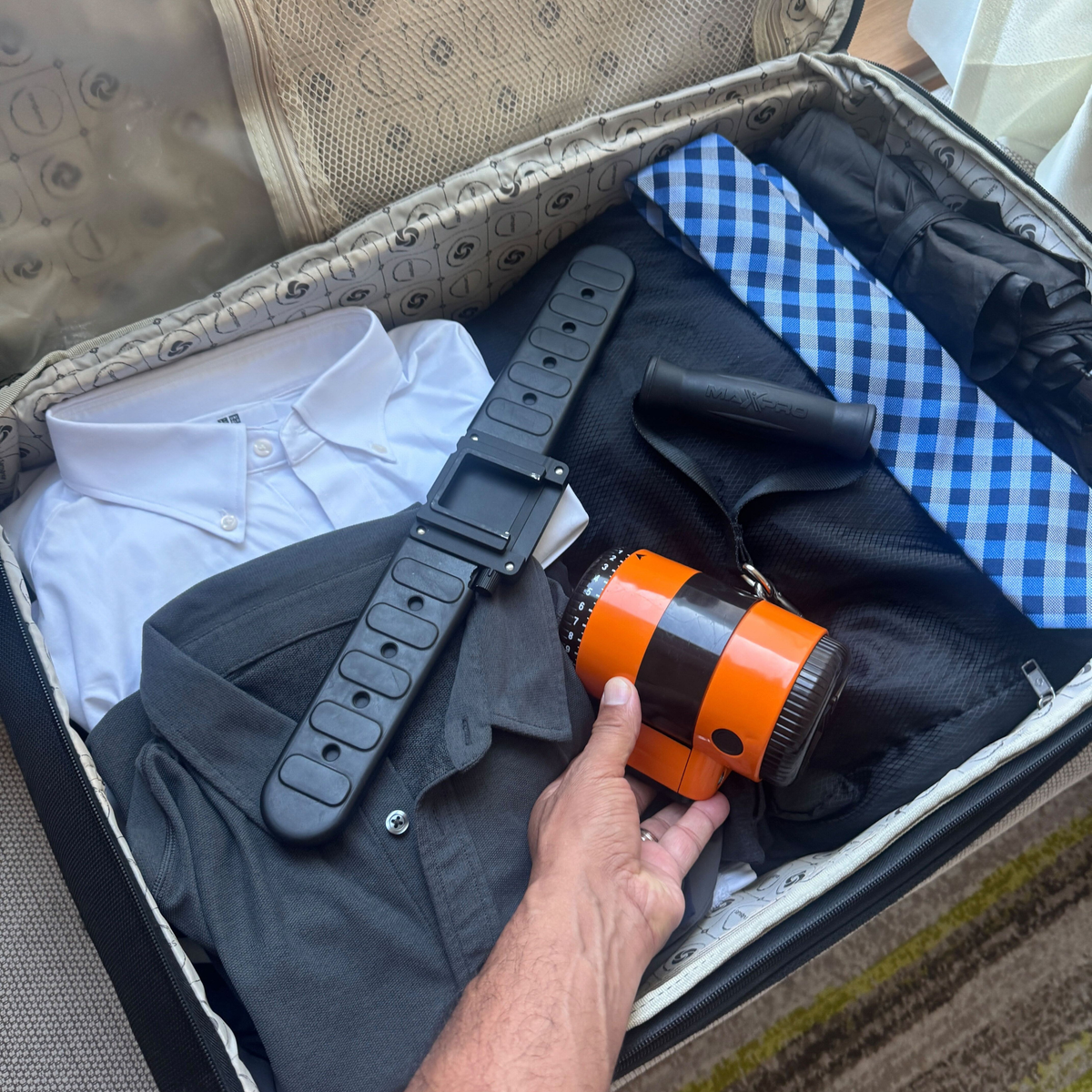 Person holding an orange and black bottle in an open suitcase with clothes and a belt.