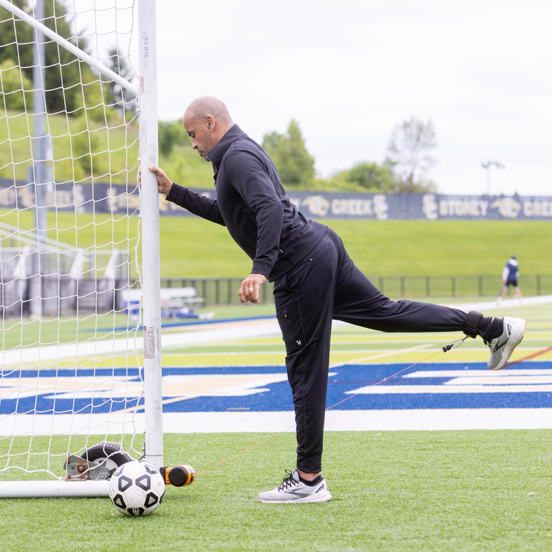 Man stretching on a soccer field with a goalpost and soccer ball in the foreground.