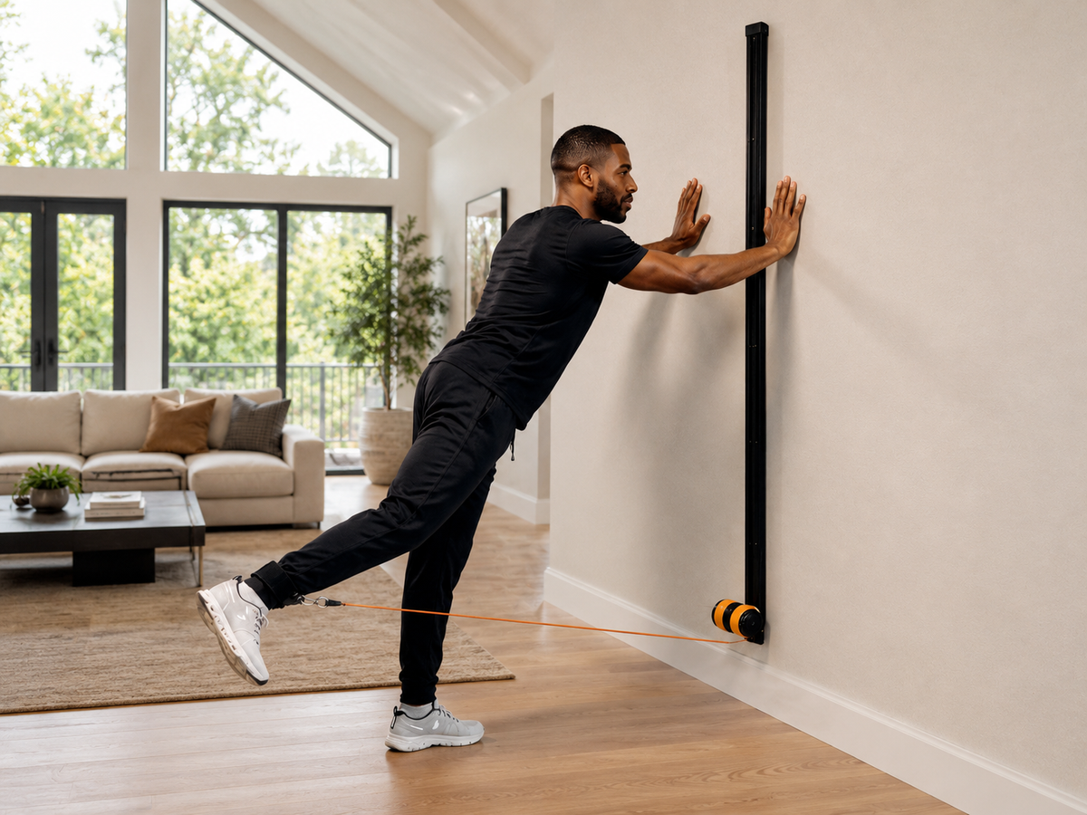 A man in a black workout outfit performs a wall push-up with a MAPRO Air attached to a black wall-mounted vertical bar in a bright, modern living room.