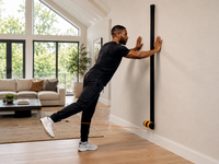 A man in a black workout outfit performs a wall push-up with a MAPRO Air attached to a black wall-mounted vertical bar in a bright, modern living room.